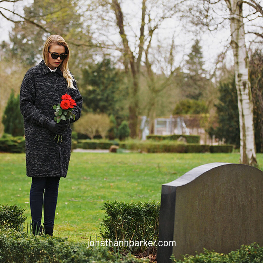 Woman standing at headstone holding red flowers in cemetary. Illustrates mourning and relates to how to navigate an estate plan after losing a loved one.